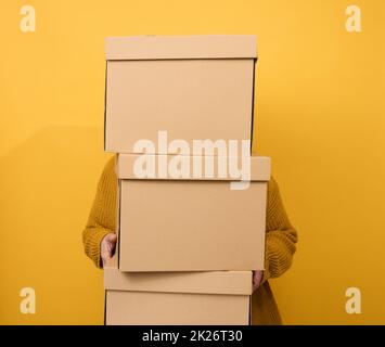 a woman in a sweater holds a stack of brown cardboard boxes on a yellow background. Relocation Stock Photo