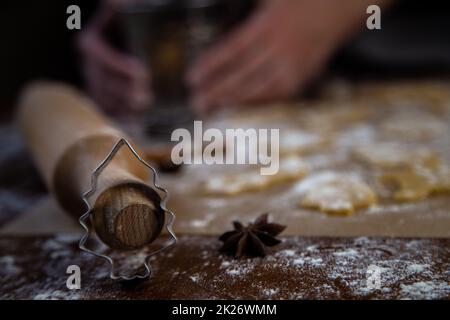 A Christmas tree-shaped mold stands leaning on a rolling pin against a background of cookies, parchment and flour cut out of dough. Photos in dark colors. Stock Photo