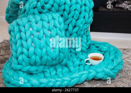 A warm plaid knitted from merino wool of a beautiful aquamarine shade lies near the fireplace along with a cup of tea. The concept of comfort. Stock Photo