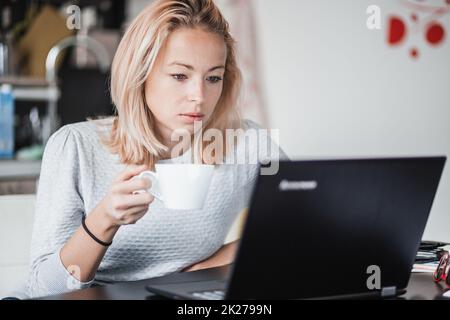 Female freelancer in her casual home clothing working remotly from her dining table in the morning. Home kitchen in the background. Stock Photo