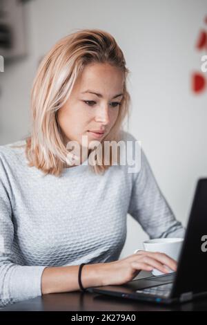 Female freelancer in her casual home clothing working remotly from her dining table in the morning. Home kitchen in the background. Stock Photo