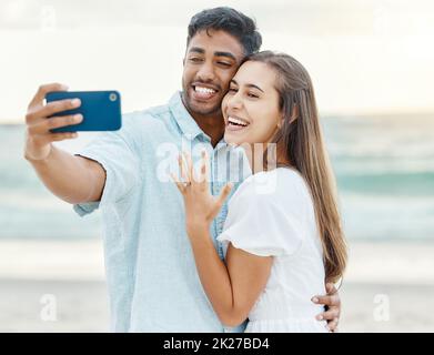 Love, beach and couple after their engagement taking a selfie on a phone while on summer vacation. Happy woman showing off her ring after a proposal Stock Photo