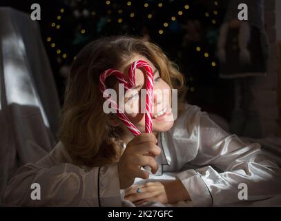 pretty young woman holds in front of face two large red lollipops folded in shape of heart, smiles cheerfully, lies under Christmas tree with garlands Stock Photo