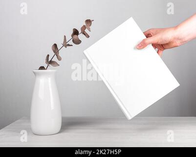 Hand holding book mockup near flowers in vase at table. Minimalist style. Reading leisure, harmony concept. Novel with white cover. High quality photo Stock Photo