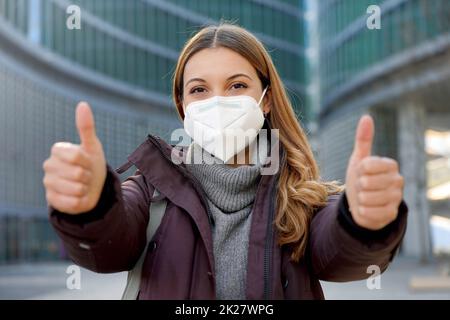 Portrait of optimistic girl wearing protective face mask showing thumbs up in modern city street and looking at camera Stock Photo