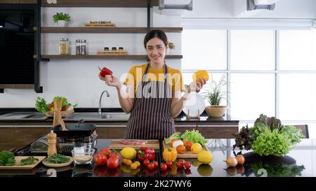 Young housewife stand smiling,  hold red and yellow bell pepper with both hands. The kitchen counter full of various kinds of vegetables. Stock Photo