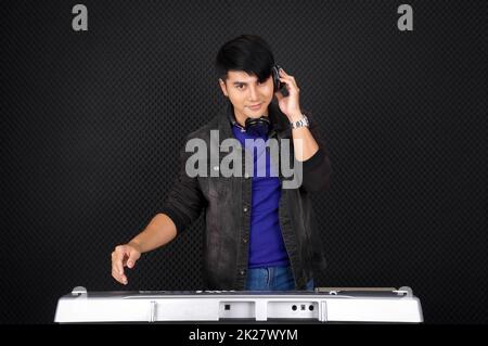 Young asian man with headphones playing an electric keyboard in front of black soundproofing wall. Musician producing music in professional recording studio. Stock Photo