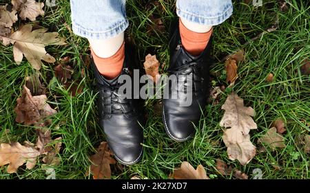 Female legs in black boots on green grass with yellow fallen autumn leaves in the park. Autumn concept. Womens shoes in autumn foliage Stock Photo