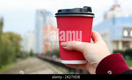 A young girl holds a red disposable paper cup with coffee or other hot drink in her hands during the cold season. Blurred street in the background. To go, away. No ecology. Stock Photo
