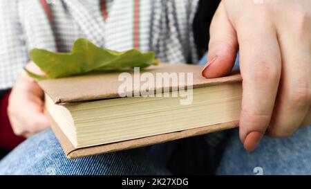 A woman holds a closed book lying in her lap with a fallen oak leaf close-up in a park on a sunny warm autumn day. The concept of relaxation, reading and relaxation alone. Stock Photo