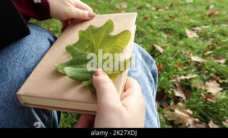 A woman holds a closed book lying in her lap with a fallen oak leaf close-up in a park on a sunny warm autumn day. The concept of relaxation, reading and relaxation alone. Stock Photo
