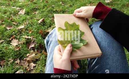 A woman holds a closed book lying in her lap with a fallen oak leaf close-up in a park on a sunny warm autumn day. The concept of relaxation, reading and relaxation alone. Stock Photo