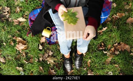 A woman holds a closed book lying on her lap with a fallen oak leaf in a park on a sunny warm autumn day on a green meadow. The concept of relaxation, reading and relaxation alone. Top view, flat lay. Stock Photo