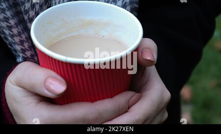 A young girl in a coat holds morning coffee with her while walking in the park. Hand holding paper cup of coffee in green park. Takeaway cappuccino. Close-up. Stock Photo