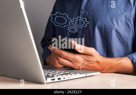 Man holding phone with virtual gears while working on laptop. Mobile settings or strategy, process organization, business idea concept. High quality photo Stock Photo