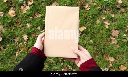 A closed book in a cover made of craft paper in female hands with green grass and fallen yellow leaves in the background. Flat lay, top view. Template, layout. Copy space. Stock Photo