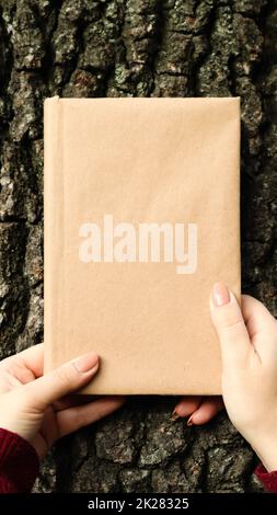 A closed book in a cover made of craft paper in female hands on the background of a tree bark in the park. Copy space. The concept of reading, recreation and leisure, study and education. Stock Photo