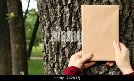 A closed book in a cover made of craft paper in female hands on the background of a tree bark in the park. Copy space. The concept of reading, recreation and leisure, study and education. Stock Photo