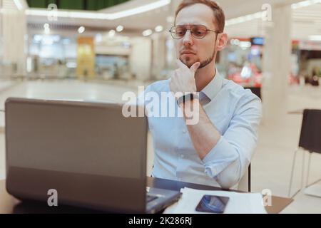 Pensive young Caucasian man sit at table food court work online on laptop pondering thinking Stock Photo