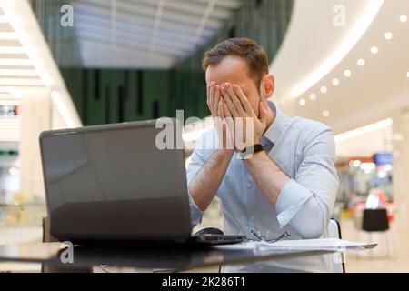 Feeling tired. Frustrated young man covering his face with hands while working on laptop Stock Photo