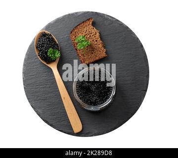 black paddlefish caviar in a wooden spoon, a slice of bread and a jar on a black round graphite board Stock Photo