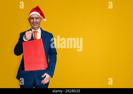 A well-dressed adult caucasian smiling male wearing a blue suit, white shirt, orange necktie and a red santa hat is holding a red paper shopping bag. Stock Photo