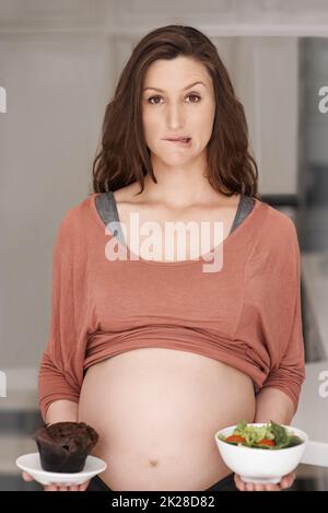I know what I SHOULD do...Cropped portrait of a young pregnant woman deciding between a salad or a cupcake in the kitchen. Stock Photo