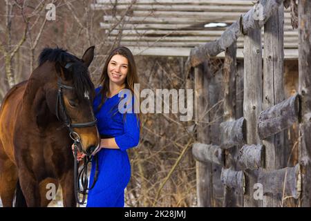 A beautiful young girl in a blue dress hugs a horse against the background of an old fence and a winter forest Stock Photo