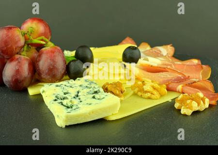 Assorted cheeses on black slate board. Brie cheese, cheddar slices,  walnuts, grapes, olives and Schwarzwald ham. Still life food. Copy space Stock Photo