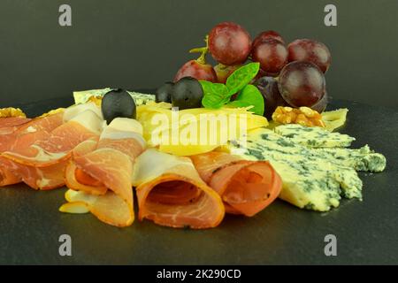 Tray with Schwarzwald ham, cheeses, walnuts, black olives. Decorated with basil. Food on slate backgroud. Copy space Stock Photo
