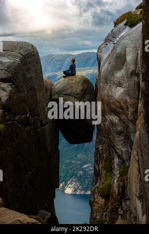 Visiting Norway Kjeragbolten located south of Lysefjorden Young woman sits on top of gian boulder in meditative pose. Stock Photo