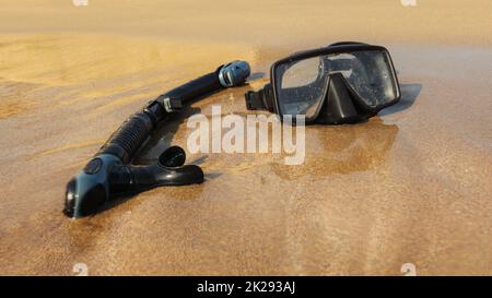Black snorkel and diving goggles on fine sandy beach. Shot through wet lens to emphasise water. Stock Photo