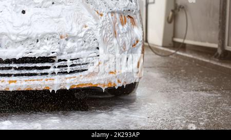 Front bumper of yellow car being washed in carwash, completely covered in white shampoo and foam, some dropping to the ground. Stock Photo