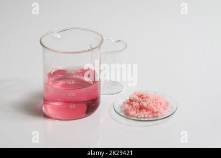 Close up inorganic chemical on white laboratory table. Pink flake chemicals in Chemical Watch Glass place next to Red EPOXY liquid rasin in beaker. Stock Photo
