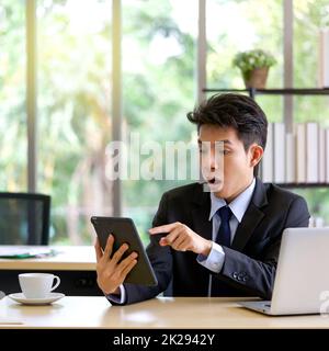 Asian  businessman in suit looking at report excited by good news, personal working with tablet computer in the office. Stock Photo