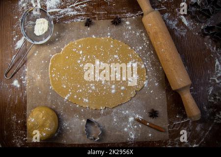 There is a rolled dough on the parchment, next to it is a lump of dough, a rolling pin, cookie cutters, cinnamon, everything is strewn with flour on a dark background Stock Photo