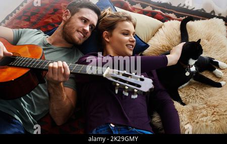 And kitty makes three. a young man playing guitar while lying on the floor with his girlfriend. Stock Photo