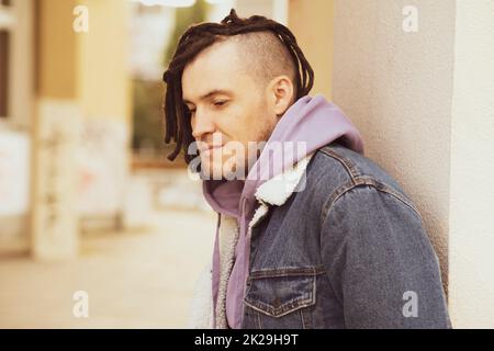 Portrait of young handsome man with dreadlocks posing, standing, leaning against wall on street Stock Photo