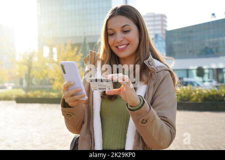 Portrait of young woman typing number of credit card on mobile phone makes easy purchase online in city street Stock Photo