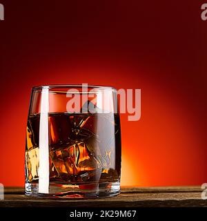 A glass of whiskey or cognac and ice cubes, close-up on a wooden table. Red background with gradient. Stock Photo