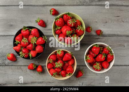 Tabletop view - strawberries in small ceramic bowls, some of them spilled on gray wood desk. Stock Photo