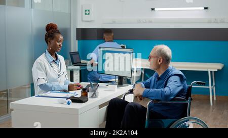 General practitioner doing checkup visit with patient in wheelchair, consulting old man with chronic disability. Medic doing examination with wheelchair user suffering from impairment. Stock Photo