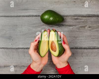 Tabletop view, young woman hands holding two halves of avocado cut in half, over gray wood desk, whole green pear next to it. Stock Photo