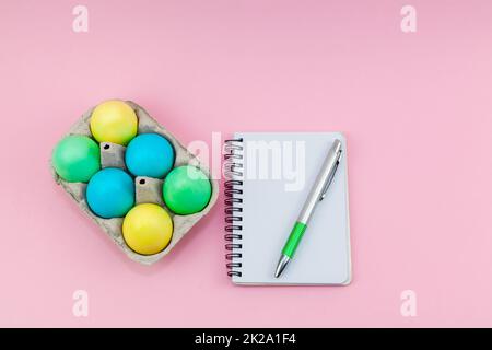 Blank notepad and pen on trendy dark pink background. Tray with multi-colored Easter eggs near the notebook. Stock Photo