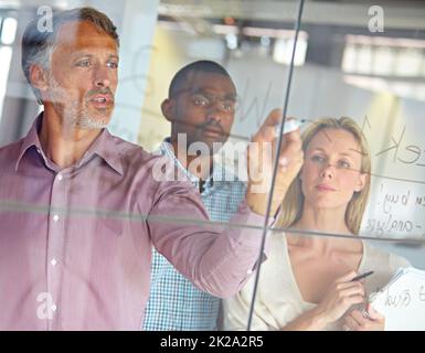 Hes a man with a plan. A mature businessman writing down plans on a glass pane while his colleagues look on. Stock Photo