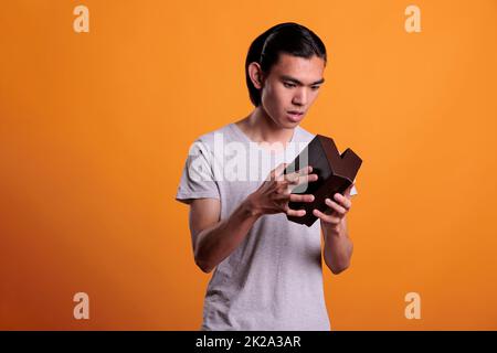 Interested asian man opening gift box, looking inside small cardboard parcel, unpacking surprise. Young person unboxing present, holding delivery package with curious facial expression Stock Photo