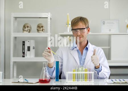 Young Brazilian scientist with a safety glasses lab is mixing cosmetic chemicals together carefully. Working atmosphere in chemical laboratory. Test tubes filled with chemicals on the table. Stock Photo