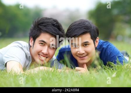 They share a love of the outdoors. Cute young gay Asian couple smiling together while lying on the grass. Stock Photo