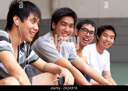 Chilling with the guys. Four young asian friends sitting in a row and smiling at the camera. Stock Photo