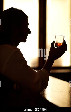 Sitting in quiet contemplation. Profile silhouette of a young man sitting at a bar and holding up a glass of whiskey. Stock Photo